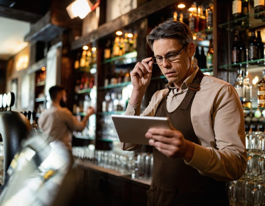 Mid adult barista using digital tablet while working in a bar. His coworker is in the background.